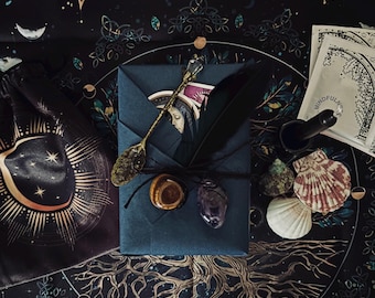 May include: An assortment of mystical items arranged on a dark, patterned surface. Includes a velvet bag with a sun and moon design, a teal envelope, a decorative spoon, crystals, a feather, seashells, and a small packet with the word "MINDFUL" printed on it.