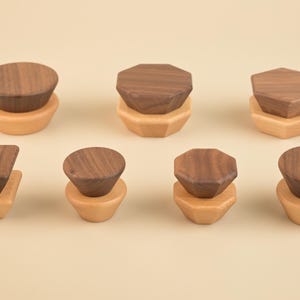 May include: A collection of eight wooden cabinet knobs in various geometric shapes. The knobs feature a dark brown top and a light brown base, set against a neutral background. The shapes include circles, squares, and octagons.