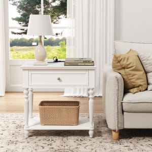 May include: A white bedside table with a drawer and a lower shelf, featuring a beige woven basket. A cream-colored lamp sits on top, with books and a black cord. The table has turned legs and is placed next to a beige sofa.