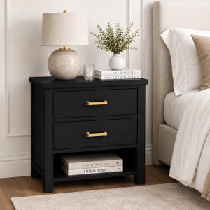 May include: Black two-drawer nightstand with gold-toned hardware. A beige lamp with a round base sits on top, along with decorative books and a small vase with greenery. A bed with white linens is visible on the right.