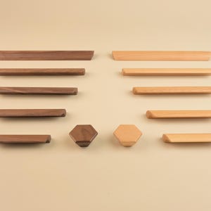 Puede incluir: Una colección de herrajes de madera para armarios, que incluye pomos y tiradores. Los herrajes tienen varias formas y tamaños, con un acabado de madera natural. La disposición incluye tiradores rectangulares, pomos redondos y pomos hexagonales.