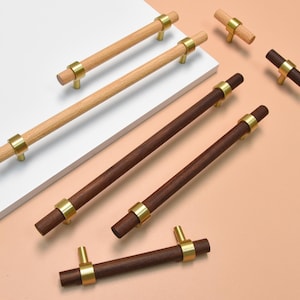 Może przedstawiać: Kolekcja drewnianych uchwytów i gałek do szafek. Uchwyty są wykonane z jasnego i ciemnego drewna z mosiężnymi metalowymi akcentami. Gałki mają podobny design. Przedmioty są ułożone na białej i brzoskwiniowej powierzchni.