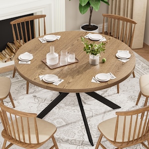 May include: A round wooden dining table with a light brown finish, set for six people. The table has a black metal base and is surrounded by wooden chairs with beige seats. The table is set with white plates, silverware, and glassware.