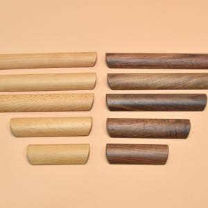 May include: A selection of wooden drawer pulls in different sizes and shades. The pulls are arranged in two columns, with lighter wood on the left and darker wood on the right. The pulls have a rounded shape and are set against a neutral background.