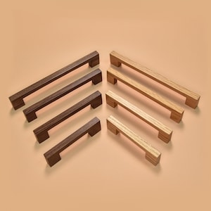 Può includere: Una collezione di maniglie per mobili in legno di varie dimensioni e tonalità di marrone e beige. Le maniglie sono rettangolari con un design semplice e moderno. La venatura del legno è visibile sulle maniglie più chiare. Sono disposte su uno sfondo neutro.