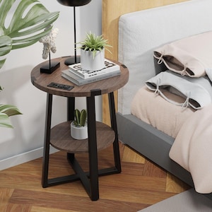 May include: A round, two-tiered side table with a dark brown wood finish and black metal legs. The top shelf holds a potted plant, books, and a small electronic device. The lower shelf features a small cactus in a white pot. The table is next to a bed.
