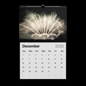 May include: A 2026 wall calendar featuring a vibrant fireworks display against a dark night sky. The December page is displayed, with a grid of dates and days of the week. The calendar is bound with a black spiral.