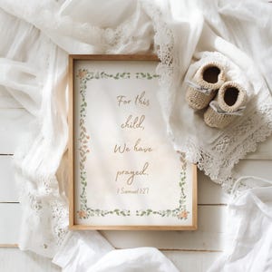 Può includere: Stampa incorniciata con il testo "For this child, We have prayed. 1 Samuel 1:27" e decorazioni floreali. Accanto, due scarpine da neonato beige a maglia con nastri grigi, su un tessuto bianco bordato di pizzo.