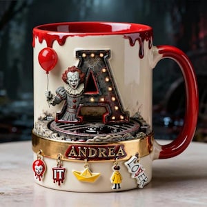 May include: Cream-colored ceramic mug with a red interior and handle. Features a 3D design of a clown holding a red balloon next to a light-up letter "A". The mug has the name "Andrea" and several dangling charms.