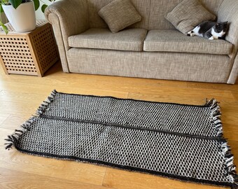 May include: A rectangular black and white woven rug with a checkered pattern. The rug has a black border and fringe on the short ends. The rug is on a wooden floor.