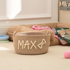 Peut inclure: Un panier à jouets pour chien tissé de couleur beige avec le nom "MAX" et un motif d'os. Le panier a deux poignées. Une balle rose et un lit pour chien sont en arrière-plan. D'autres accessoires pour chiens sont également visibles.