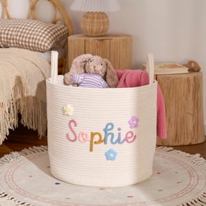 Könnte beinhalten: Ein weißer Seilkorb mit dem Namen "Sophie" in bunten Fäden gestickt, verziert mit Blumenakzenten. Der Korb enthält einen Stoffhasen und eine rosa Decke. Der Korb steht auf einem runden Teppich mit Fransen.