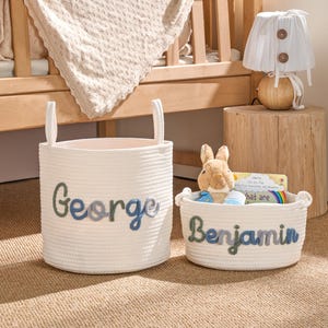 May include: Two white rope baskets, one large and one small, with the names "George" and "Benjamin" stitched in blue and green. A plush bunny and a book are in the smaller basket. A wooden crib and lamp are in the background.