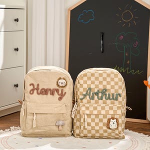 May include: Two beige backpacks, one corduroy and one checkered, are shown. The corduroy backpack has the name "Henry" and a lion patch, the checkered one has "Arthur" and a hedgehog patch. A chalkboard with drawings is in the background.