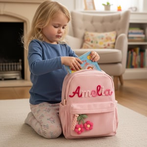 Könnte beinhalten: Ein rosa Cord-Rucksack mit dem Namen "Amelia" in roter Schrift gestickt. Der Rucksack hat eine Vordertasche und ein Kirschdesign. Ein Kind legt ein Buch in den Rucksack.