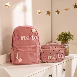 Könnte beinhalten: Ein rosa Cord-Rucksack und eine passende Lunchtasche, beide mit dem Namen "Amelia" in einer weichen, erhabenen Schrift personalisiert. Der Rucksack hat eine Vordertasche und eine dekorative Blume. Die Lunchtasche hat ein Erdbeermuster.