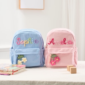 May include: Two corduroy backpacks, one light blue and one pink, are displayed. The blue backpack has the name "Sophie" in rainbow thread, and the pink backpack has the name "Amelia" in pink thread. Both have front pockets and floral embellishments.