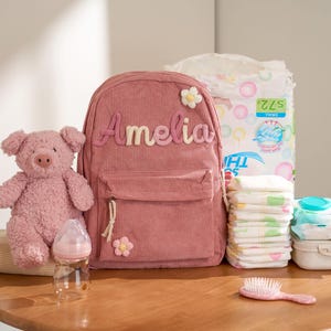 May include: A pink corduroy backpack with the name "Amelia", a stuffed pig, a baby bottle, nappies, wipes, and a hairbrush. The backpack has a front pocket and a flower applique. The items are arranged on a wooden surface.