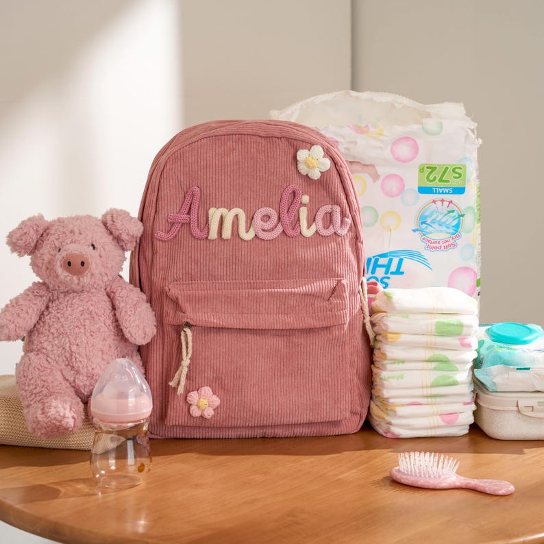 May include: A pink corduroy backpack with the name "Amelia" in a script font, a stuffed pig, a baby bottle, diapers, wipes, and a hairbrush. The backpack has a front pocket and a flower applique.