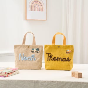 May include: Two corduroy tote bags, one tan and one yellow, with embroidered names and small vehicle patches. The tan bag has "Noah" in blue script and a camper van. The yellow bag has "Thomas" in brown script and a construction vehicle.