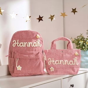May include: A pink corduroy backpack and tote bag set. The backpack has a front pocket and the name "Hannah" in white script. The tote bag also features the name "Hannah" and flower and bee embellishments.