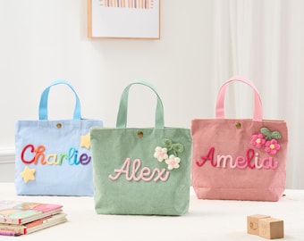 May include: Three corduroy tote bags in blue, green, and pink, each with a name embroidered on the front. The blue bag says "Charlie" in rainbow colors, the green bag says "Alex", and the pink bag says "Amelia". Each bag has a matching handle.