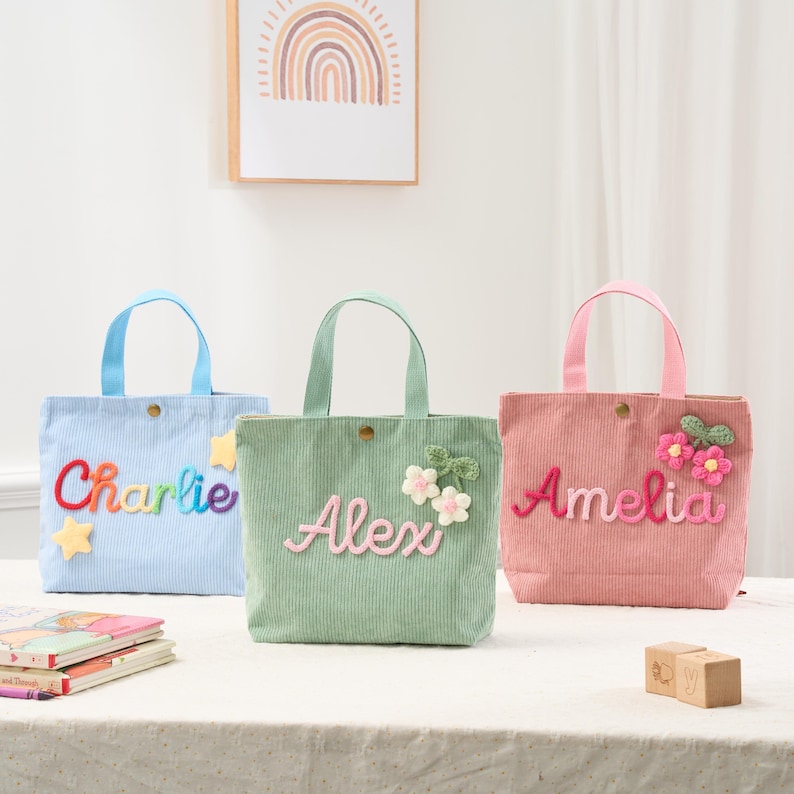 May include: Three corduroy tote bags in blue, green, and pink, each with a name embroidered on the front. The blue bag says "Charlie" in rainbow colors, the green bag says "Alex", and the pink bag says "Amelia". Each bag has a matching handle.