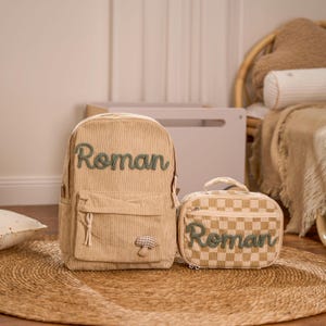 Op de afbeelding: Een beige corduroy rugzak en een bijpassende lunchtas. Beide items zijn voorzien van de naam "Roman" in groene letters. De rugzak heeft een voorvak en een paddenstoel applicatie. De lunchtas heeft een dambordpatroon.