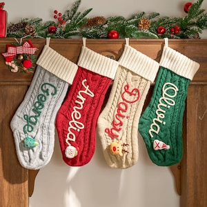 May include: Four cable-knit Christmas stockings hang from a wooden mantel. The stockings are grey, red, beige, and green, each with a white cuff and a name embroidered in a contrasting colour. Decorative accents include a snowman, reindeer, and a Santa hat.