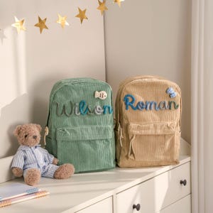 Puede incluir: Dos mochilas de pana, una verde y otra beige, están una al lado de la otra. La mochila verde tiene el nombre "Wilson" en escritura azul, y la mochila beige tiene el nombre "Roman" en escritura azul. Un oso de peluche está delante de las mochilas.