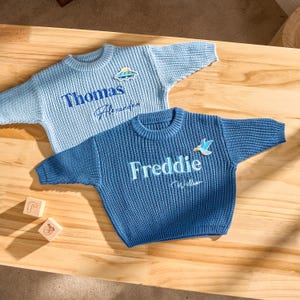 May include: Two blue knitted sweaters with embroidered names and small patches. One is light blue with "Thomas Alexander" and a UFO patch. The other is a darker blue with "Freddie Wilson" and a bird patch. Wooden blocks are in the foreground.
