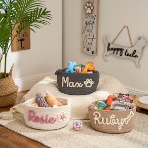 May include: Three woven pet toy baskets, each with a pet's name embroidered on the front. The gray basket says "Max" and has a paw print. The white basket says "Rosie" with a flower. The brown basket says "Ruby" with a bone.