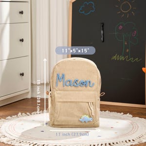May include: Beige corduroy backpack, 11" x 5" x 15", with the name "Mason" in blue letters. Features a small dinosaur patch. The backpack is 15 inches (38.1 cm) tall and 11 inches (27.9 cm) wide.