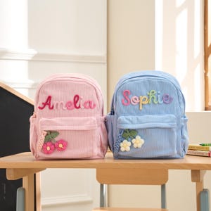 May include: Two corduroy backpacks, one pink and one light blue, sit on a wooden table. The pink backpack has the name "Amelia" embroidered in pink, and the blue backpack has the name "Sophie" embroidered in rainbow colors. Both have floral accents.