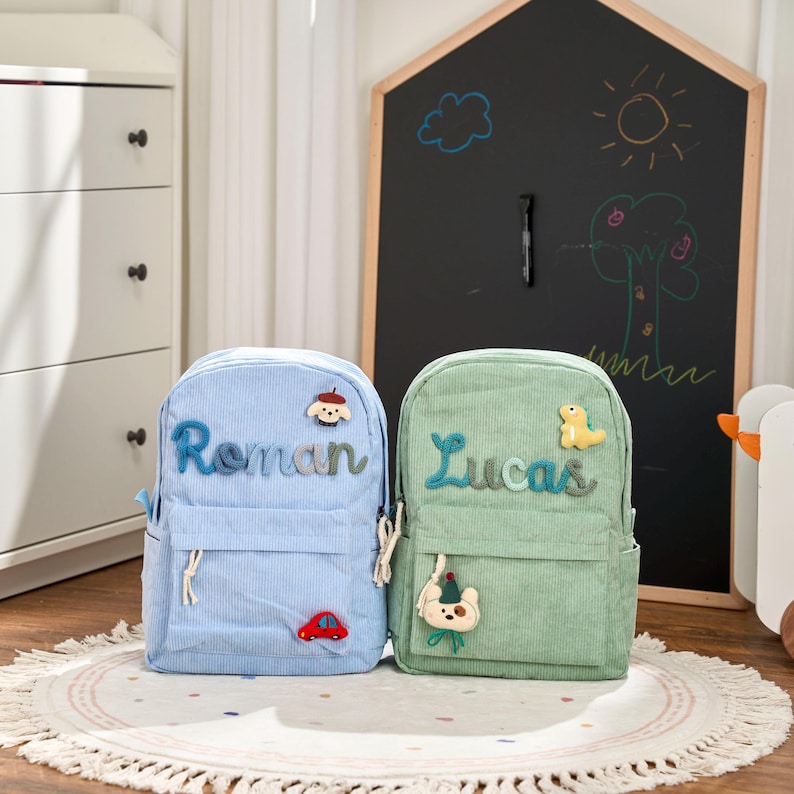 May include: Two corduroy backpacks, one light blue and one green, each personalized with a name in blue and green yarn, respectively. The blue backpack has a red car and a cloud patch. The green backpack has a yellow dinosaur and a bear patch.