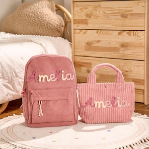 May include: A pink corduroy backpack and tote bag set. The backpack has a front pocket and the name "Amelia" embroidered in a cursive font. The tote bag also features the name "Amelia". Both items are on a round rug.