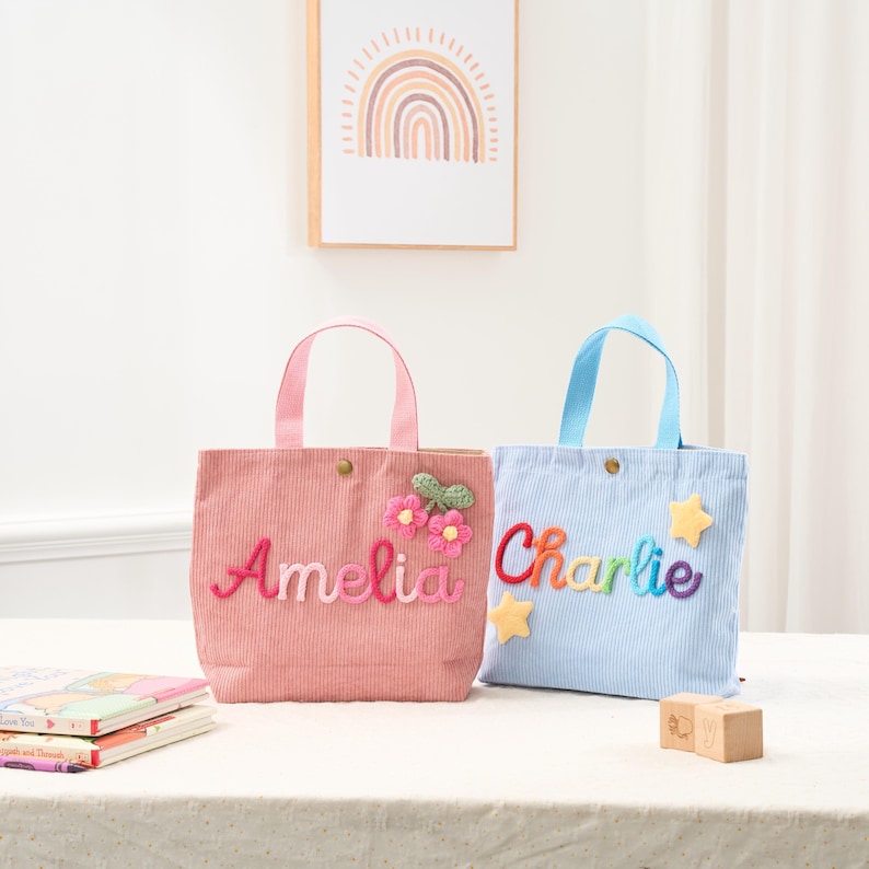 May include: Two corduroy tote bags, one pink and one blue, with embroidered names and floral or star embellishments. The pink bag says "Amelia" and the blue bag says "Charlie". Both have matching handles.