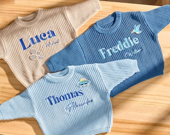 May include: Three knitted jumpers in beige, blue, and light blue, each embroidered with a name in a contrasting colour. The jumpers have patches with small designs. The names are Luca, Freddie, and Thomas.