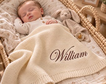 May include: A cream-coloured knitted baby blanket featuring the name William embroidered in dark brown script. The blanket is draped over a sleeping infant wearing a white long-sleeved shirt with buttons, alongside a plush bunny toy.