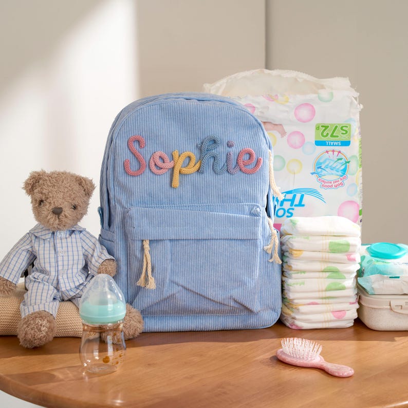 May include: A light blue corduroy backpack with the name "Sophie" in colorful letters, a teddy bear in pajamas, a baby bottle, diapers, wipes, and a pink hairbrush. The backpack has a front pocket and tassel details.