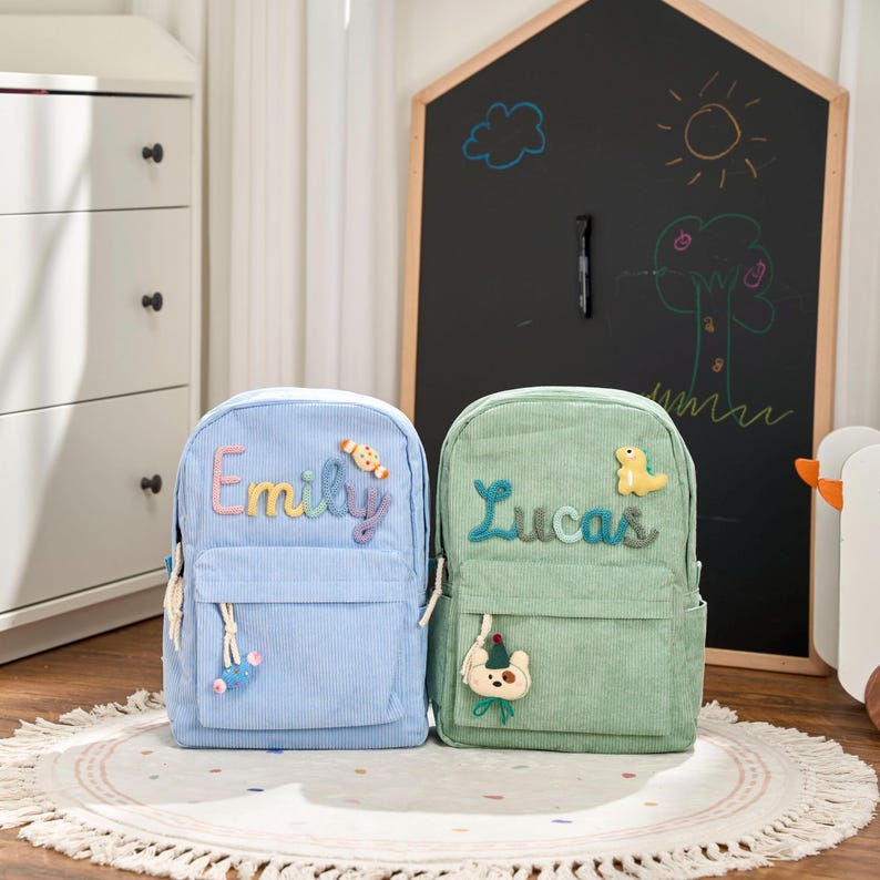 May include: Two corduroy backpacks, one light blue and one green, each with a front pocket and a name embroidered on the front. The blue backpack says "Emily" in rainbow thread, and the green backpack says "Lucas". Both have decorative charms.