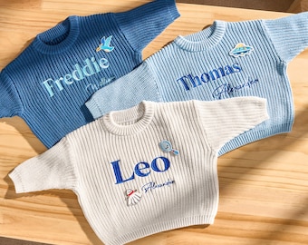 May include: Three personalised knitted sweaters in shades of blue and white. Each sweater has a name embroidered in a contrasting colour, plus a small decorative patch. The sweaters are displayed on a wooden surface.