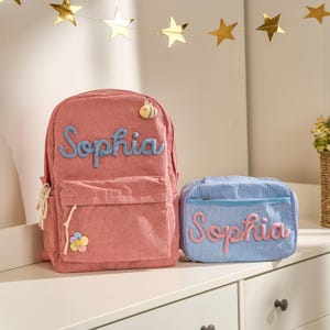 Op de afbeelding: Een roze corduroy rugzak en een lichtblauwe lunchtas, beide met de naam "Sophia" in script. De rugzak heeft een voorvak, een bijenapplicatie en een bloem. De lunchtas is rechthoekig.