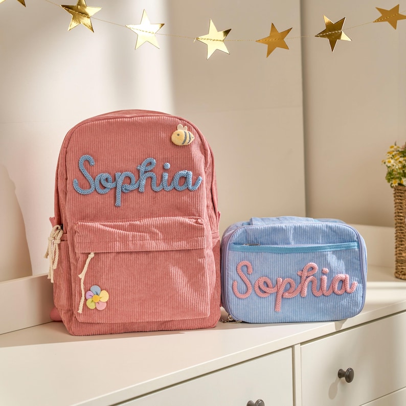 Op de afbeelding: Een roze corduroy rugzak en een lichtblauwe lunchtas, beide met de naam "Sophia" in script. De rugzak heeft een voorvak, een bijenapplicatie en een bloem. De lunchtas is rechthoekig.
