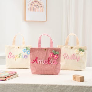 May include: Three corduroy tote bags in cream and pink, each with a name embroidered in colorful thread. The bags feature floral embellishments and matching handles. The bag on the left says "Sophie", the middle bag says "Amelia", and the right bag says "Ruby".