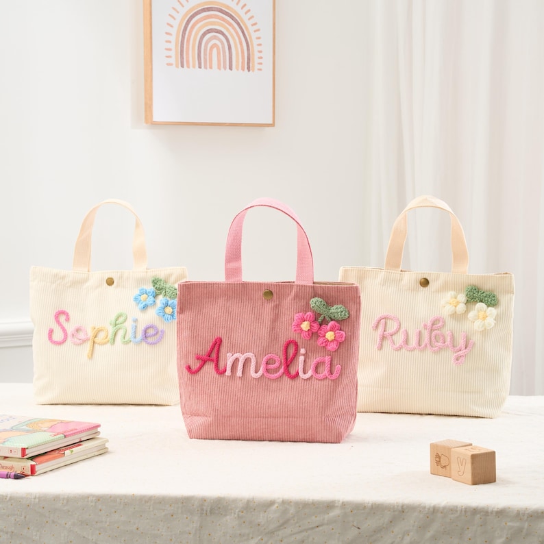 May include: Three corduroy tote bags in cream and pink, each with a name embroidered in colorful thread. The bags feature floral embellishments and matching handles. The bag on the left says "Sophie", the middle bag says "Amelia", and the right bag says "Ruby".