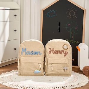 May include: Two beige corduroy backpacks with personalized names, "Mason" in blue and "Henry" in brown. Each backpack has a front pocket and decorative patches, including a dinosaur and a lion. The backpacks are on a round rug.