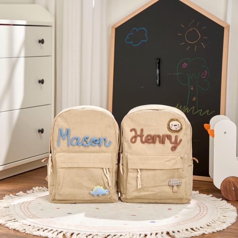 May include: Two beige corduroy backpacks with personalized names, "Mason" in blue and "Henry" in brown. Each backpack has a front pocket and decorative patches, including a dinosaur and a lion. The backpacks are on a round rug.
