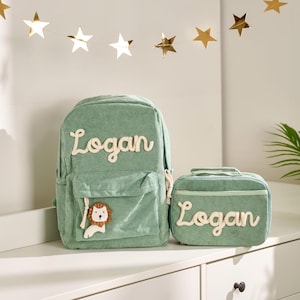 Könnte beinhalten: Ein salbeigrüner Cord-Rucksack und eine passende Lunchtasche. Beide Artikel tragen den Namen "Logan" in cremefarbenen Buchstaben. Der Rucksack hat eine Vordertasche und eine Löwenapplikation. Goldene Sternengirlande im Hintergrund.