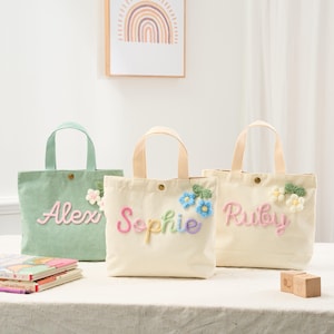 May include: Three corduroy tote bags in pastel colors, each with a name embroidered in colorful thread. The bags have matching handles and floral accents. The names are Alex, Sophie, and Ruby.